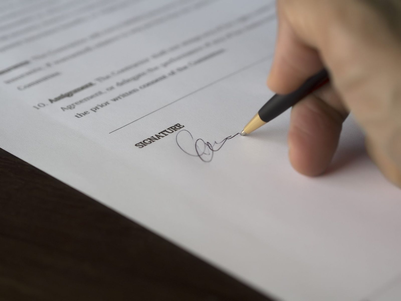Mão assinando um documento com caneta sobre uma mesa de madeira